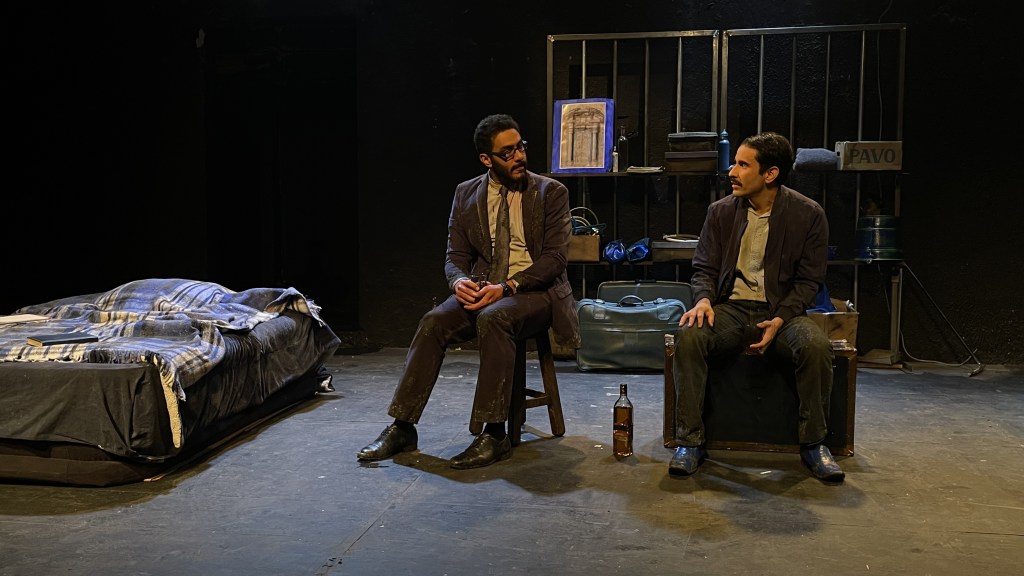 Jesús Delgado y Sebastián Torres comparten escena en "Emigrantes". Jesús Delgado viste ropa haraposa y se sienta en una maleta oscura mientras que Sebastián Torres viste un traje sastre sucio y corbata desgastada mientras se sienta en un banco de madera.